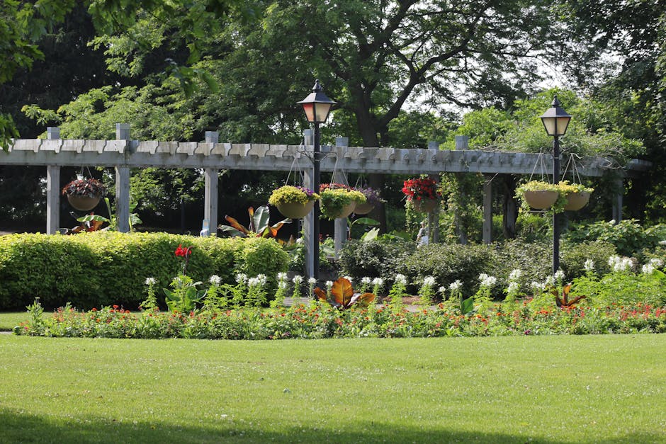 A well-maintained garden scene featuring a lush, neatly mowed lawn in the foreground with a border of colorful flowering plants and shrubs. Behind the flower border, a trimmed hedge runs horizontally 